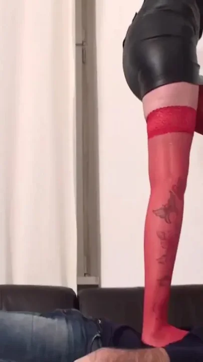 Tattoed redhead in leather skirt and red stockings crushing slave on leather couch